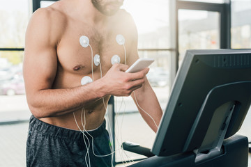 cropped view of sportsman with smartphone running on treadmill during endurance test in gym