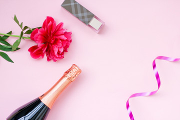 festive layout girls, a bottle of champagne, cosmetics, red flower peony on a pink background. place for text, top view.