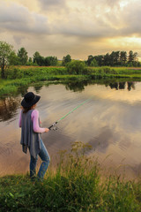 girl in a hat fishing rod on the lake, summer landscape, storm clouds in the sky. outdoor activities. rear view