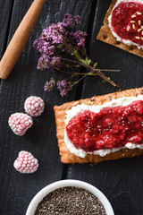 Superfood breakfast flat lay. Chia seed jam with raspberries and ricotta on rye crispbread with marjoram flowers on black background