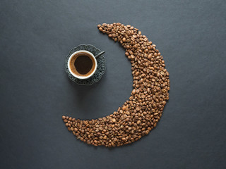Crescent shape made of coffee beans and a cup of black coffee on black background. Top view