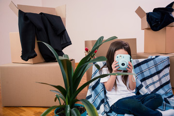 Happy cute preschooler girl sitting in living room on a blanket excited to move to new flat, is taking photo on camera