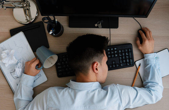 Top view of a young specialist that fell asleep at the workplace in the office in front of the monitor and knocked over a mug. Overwork concept.