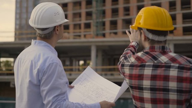 Two architects analyze blueprint of building standing indoors. They hold construction documents in their hands and point with pen on details depicted on paper, talk each other and analyze information