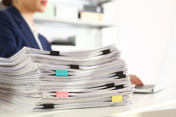 Stack of documents and woman working with laptop at table in office, closeup