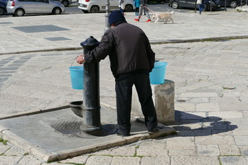 Anziano signore in piazza alla fontana mentre riempie d'acqua due secchi di plastica