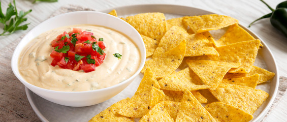 Homemade cheesy dip in a bowl with yellow tortilla chips on a white wooden surface, low angle view....