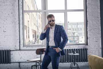 Looking perfect. Full length of handsome young bearded man in full suit and sunglasses looking to the camera while speaking by his cell phone. standing at loft interior