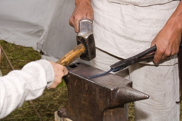 A blacksmith teaches a boy working with a hammer. Forging metal products on the anvil in the open...