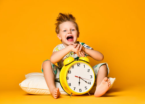 Little cute boy in pajamas holding a toy dinosaur in his hands, sitting on a pillow with an alarm clock. Isolated on a yellow background.