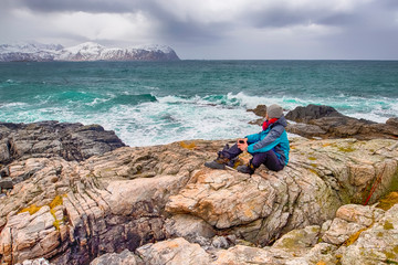 Young Man in Vivid Green Jacket Sitting on Mountain and Drinking Tea. Against Snowy Summits.
