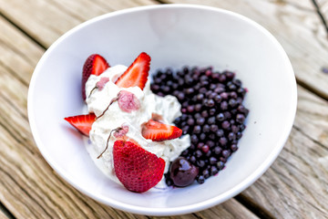 Closeup of colorful bowl of vanilla ice cream topped with fresh cut slices of red strawberry large berries, blueberries with sakura cherry blossom flowers pink petals