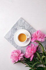 Fashion blogger styled desk frame with flowers, cup of herbal tea, pink peonies on marble plate on white, breakfast morning lifestyle, minimal concept, copy space