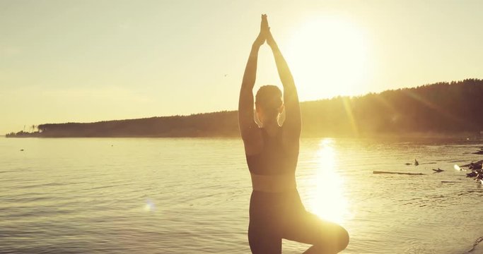 Young woman is practicing yoga in tree pose standing on the pier by the river. Girl is training in the yellow rays of the sunset, back view. She is keeping balance on one leg.