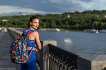 girl walks at sunset along the embankment in a blue sports suit with a colored backpack. Summer...