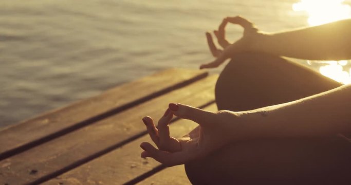Woman is meditating sitting in lotus pose, side view. She is holding hands in namaste mudra pose, hands closeup. Yellow wonderful sunset over the water.