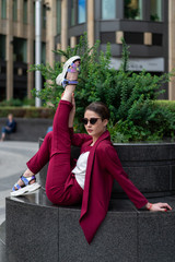 cool model with a short haircut in glasses and a business pantsuit shows stretching. Girl posing in a jacket and trousers of burgundy color outdoors in summer day