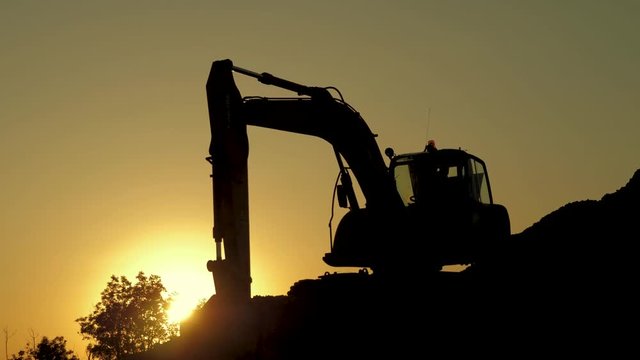Silhouette of an excavator that loads sand into a truck at sunset. Concept construction and heavy industry, machine will be used in heavy industry business. Slow motion footage.