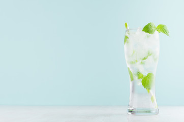 Healthy fresh tonic with green mint, ice cubes, soda, striped straw in misted glass on white wood table, pastel green color background.