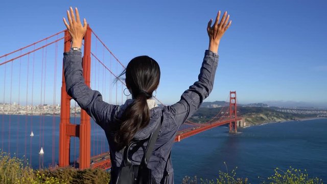 back view cheering young asian woman backpacker at seaside mountain peak raising hands with freedom crefree nature wild lover. girl hiker sightseeing red iron golden gate bridge by ocean sea sky.