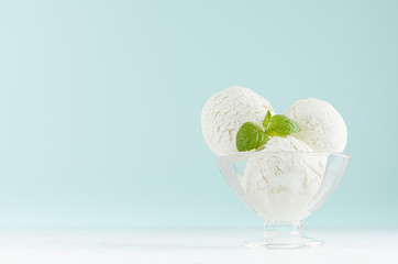 Summer cold dessert - three creamy ice cream balls in elegant glass bowl with green mint on white wood table and pastel blue wall.