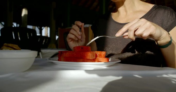 Close-up of a woman sitting at a table, she holds a fork and a knife, cuts and eats watermelon.