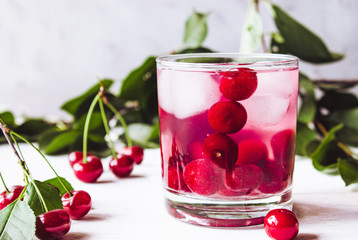 A glass of cherry drink with ice in a glass on a white wooden background. Fresh summer cocktail with cherries and ice cubes.