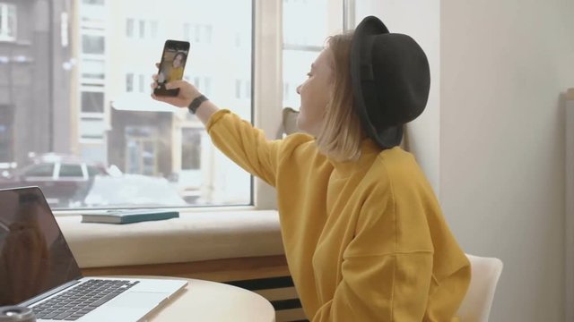 Young pretty blonde female blogger using smart phone, typing, taking selfie, chatting, while sitting at the cafe, using laptop on the table, near the window, she is wearing yellow sweater and hat