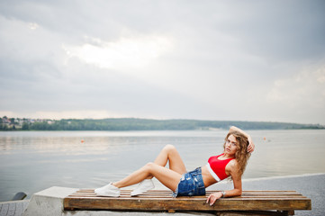 Fototapeta na wymiar Sexy curly model girl in red top, jeans denim shorts, eyeglasses and sneakers posed on bench against lake.