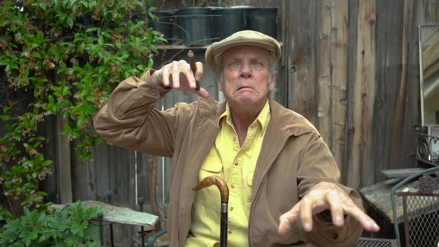 Deranged old man making crazy face and holding both hands up like claws; holding cigar in one hand. Slow motion zoom in.