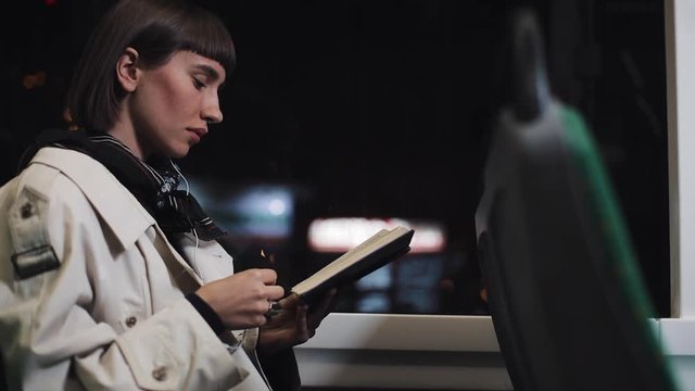 Young woman or passenger reading book sitting in public transport, steadicam shot. Slow motion. City lights background. Commuter, student, knowledge concept.