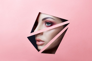 Beauty face red makeup eyes of a young girl in a slit hole of pink red paper. Woman with beautiful makeup red glowing shadow, big blue eyes in the slit hole. Advertising copy space