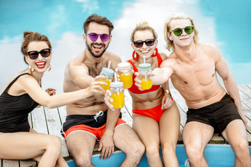 Group of happy friends sitting together by the swimming pool, talking and having fun drinking...