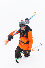 Portrait of a professional skier athlete in a knitted hat and orange-black suit with a black ski mask with skis on his shoulder during a snowstorm on a light background in the snow. Wide angle