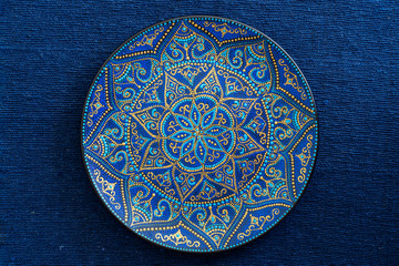 Decorative ceramic plate with blue and golden colors, painted plate on the background of blue fabric, closeup. Decorative porcelain plate painted with acrylic paints, handwork