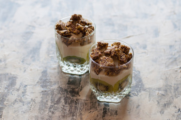 Granola, kiwi, banana and greek yogurt in glass cups on gray concrete