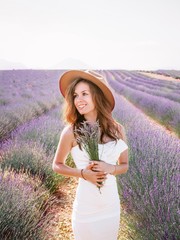 Young brunette girl with long hair in a white formal dress and a felt hat holds a bouquet of lavender and poses in a lavender field in Prvoance, France
