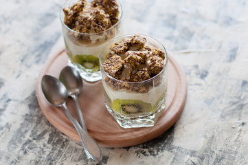 Dessert granola, Greek yogurt, kiwi and banana in two glass cups with spoons on wooden round board, gray concrete, close-up, view from the top. Fitness, figure, body and healthy food