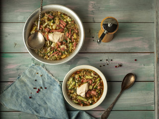 Delicious summer soup on kvass with fresh vegetables and sausage in two different plates with tablespoons and a napkin on a wooden turquoise tray, in the background a wooden pepper shaker
