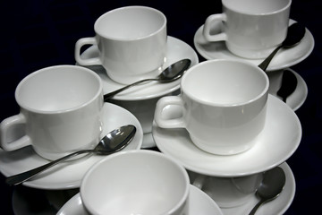 still life of rows of empy white cups and saucers with metal teaspoons side view on black background close up