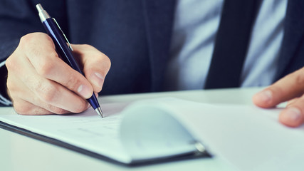 Hand of businessman in suit filling and signing with blue pen partnership agreement form clipped to pad closeup.