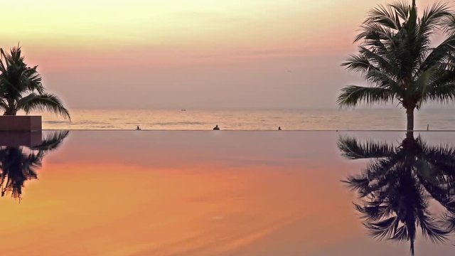 Static shot of an infinity pool during sunset, showing the orange colored sky and a silhouette of palm trees and the ocean in the background