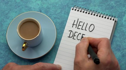 Hello December - a man hand writing a note with a black marker in a notebook, overhead view with a cup of coffee