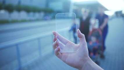 A woman in the city, uses transparent smartphone with touchscreen advanced technology. Close-up person holding invisible mobile phone in the street against people with kids walking by. - Powered by Adobe