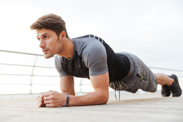 Photo of masculine man in tracksuit doing plank while working out on wooden pier at seaside
