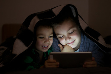 Cute little children lying under blanket with book and tablet computer. Brothers with tablet computer in a dark room smiling