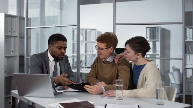 Waist-up shot of 30-something Caucasian couple sitting at table in notary office and listening to professional Afro-American middle-aged man, who is demonstrating them document and explaining terms