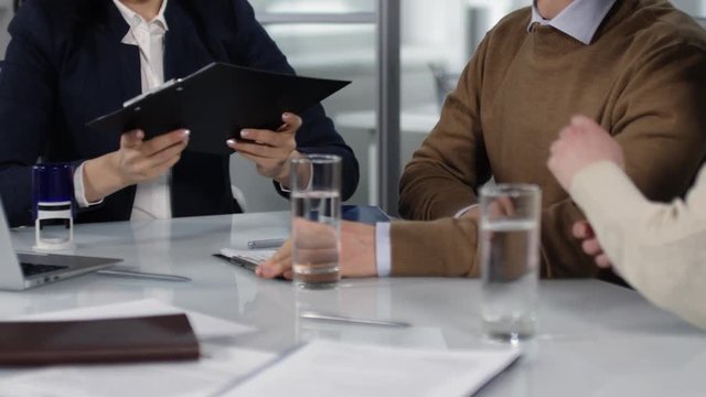 Medium body shot of man and woman sitting at table at solicitor office, handing over signed documents on clipboards to professionally dressed woman, who is then stamping them