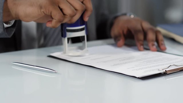 Tracking shot hands shot of dark skinned man in business suit sealing legal paperwork with round self inking rubber stamp