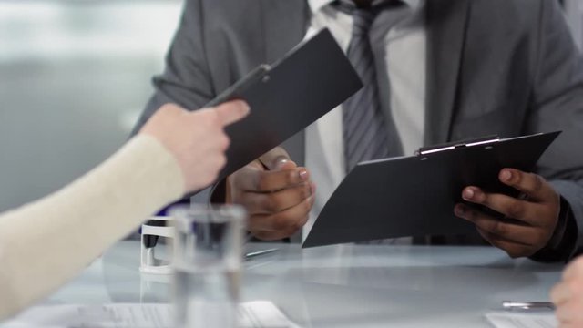 Tracking hands shot of dark-skinned male notary, dressed in business suit and tie, giving separate copies of unsigned draft contract to Caucasian male and female clients